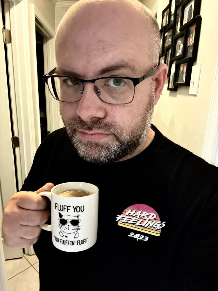 Bald, bearded man with glasses wearing a black t-shirt with an 80’s movie style font saying “Hard Feelings 2023” from Iliza Schlesinger’s live show. He’s also holding a coffee mug showing a cat with shades, flipping off with both hands, and it says “Fluff you, you fluffin’ fluff!”