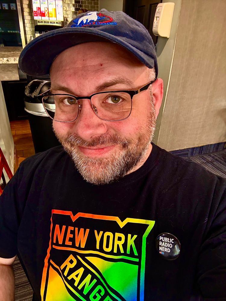Bardd wearing a blue hat and black NY Rangers (hockey team) t-shirt, with their logo in rainbow colors! 