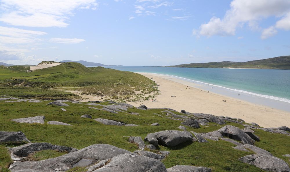 The yellow sands of Luskentyre Beach on the Isle of Harris under a blue sky. It's hard to see in the photo but there's a small graveyard on the left side in the grass behind the beach, with rows of gravestones facing off against each other across a gap in between. 