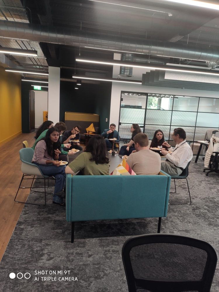 Group of people gathered in a modern office lounge, sitting on teal couches and chairs, eating and conversing. The atmosphere is casual and social.