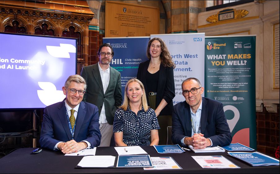 A group of five people, three seated and two standing, pose at a table with documents. Behind them, banners and a screen display health-related content. The setting is formal and professional.