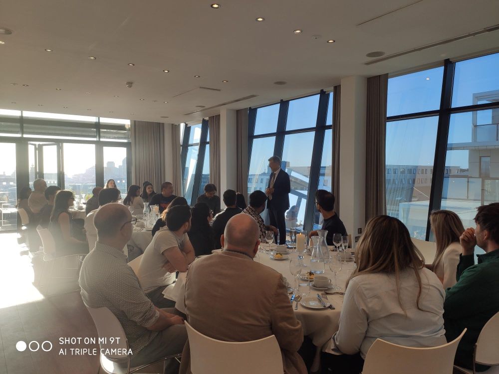 A man stands speaking to a seated group in a bright room with large windows. Attendees listen attentively around tables set with glasses and cutlery.