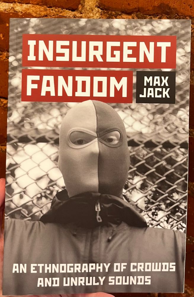Cover of Insurgent Fandom by Max Jack. A black & white cover photo shows an ultra whose face is covered by a bi-color ski mask. He stares resolutely into the camera while standing in front of a chainlink fence. 