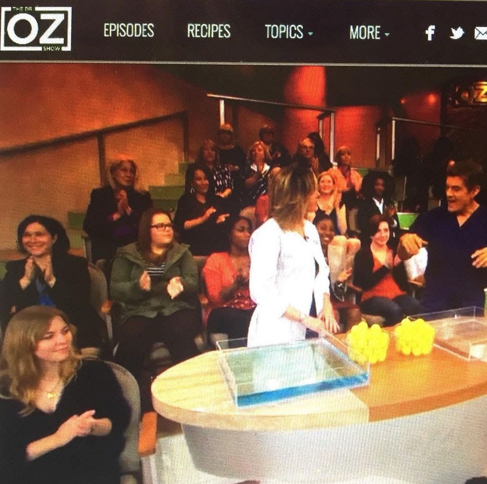 Screenshot of a live audience during a 2015 taping of the Dr. Oz Show. I am seated in the bottom left corner next to a display of ping pong balls and two plexiglass dishes, one of which is filled with blue water. During the show, a guest tossed balls into each dish. The empty dish repelled balls while the dish filled with water retained the balls, proving that skin needs moisture to retain additional moisture. The episode was sponsored by a Eucerin in-shower moisturizer. 