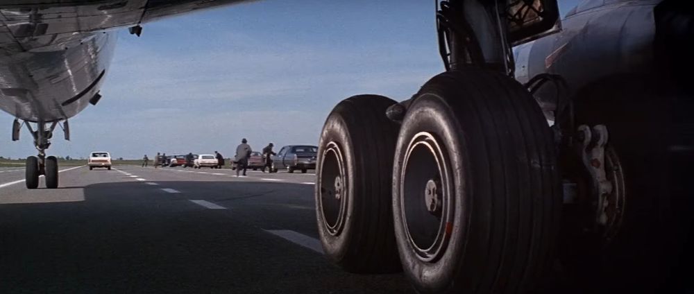 Frame from the French crime film 'Le clan des Siciliens'/'The Sicilian Clan' (1969). View from underneath a passenger airliner that has landed on a closed highway. In the foreground on the right the wheels and the landing gear from the right wing of the plane. In the background men getting into cars parked one after another on the right side of the highway.