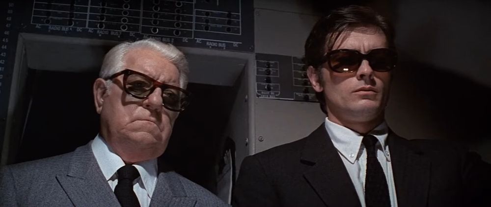 Frame from the French crime film 'Le clan des Siciliens'/'The Sicilian Clan' (1969). Close-up of two standing next to each other in the flight deck of a passenger airliner. On the left a white haired older man (Jean Gabin as Vittorio Manalese) wearing sunglasses, a grey suit and a black tie, on the right a dark haired younger man (Alain Delon as Roger Sartet) wearing sunglasses, a black suit and a tie.