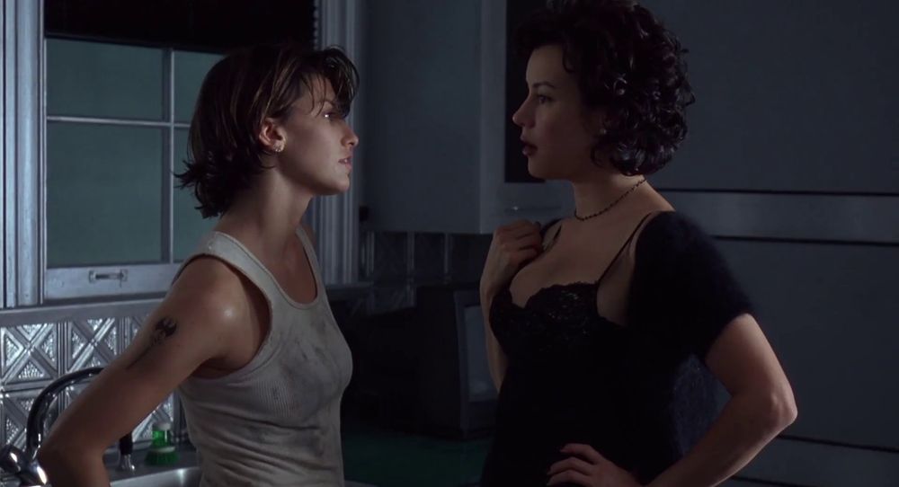 Frame from the neo-noir thriller 'Bound' (1996). Two women standing facing each other in a kitchen. On the left a woman (Gina Gershon as Corky) with short dark brown hair wearing a white dirty wifebeater and on the right a woman (Jennifer Tilly as Violet) with short black wavy hair wearing a black negligée.