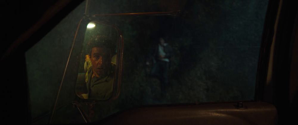 Frame from the horror film 'Wolf Man' (2025). View through the left side window of a truck that's hanging up a tree at night. There's a man lying on his back down on the ground. Dark haired man (Christopher Abbott as Blake Lovell) sitting on the driver's seat seen on the wing mirror.