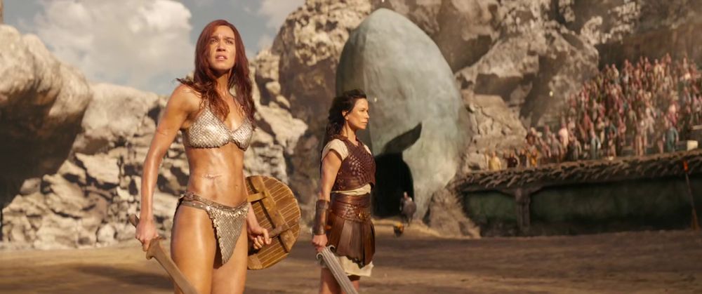 Frame from the sword and sorcery film 'Red Sonja' (2025). Ginger haired female warrior (Matilda Lutz as Red Sonja) wearing a shiny metal bikini and holding a wooden sword in her right hand and a round wooden shield on her left standing on a gladiatorial arena next to another female warrior (Rhona Mitra as Petra) with dark brown hair wearing a brown leather skirt and top on top of a white dress holding a sword in her right hand.
