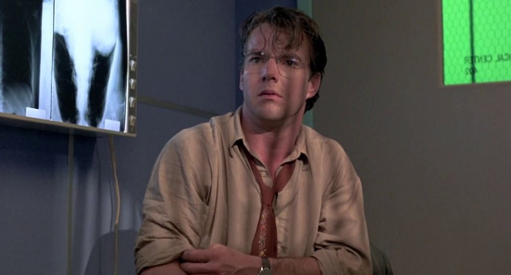Frame from the neo-noir thriller 'D.O.A.' (1988). A spectacled brown haired man (Dennis Quaid as Prof. Dexter Cornell) wearing a khaki shirt and a maroon tie sitting in a hospital room next to an X-ray viewer on the wall on the left.