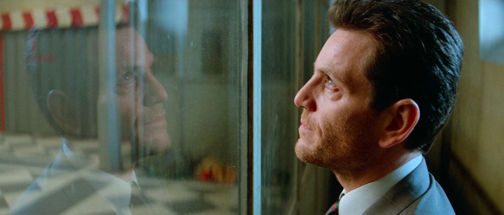 Frame from the French action thriller 'Nikita'/'La Femme Nikita' (1990). Close-up of a brown haired man (Tchéky Karyo as Bob) with stubbled beard wearing a grey suit, a tie and a white shirt standing next to a glass wall (on the right). His face can be seen reflected on the glass.