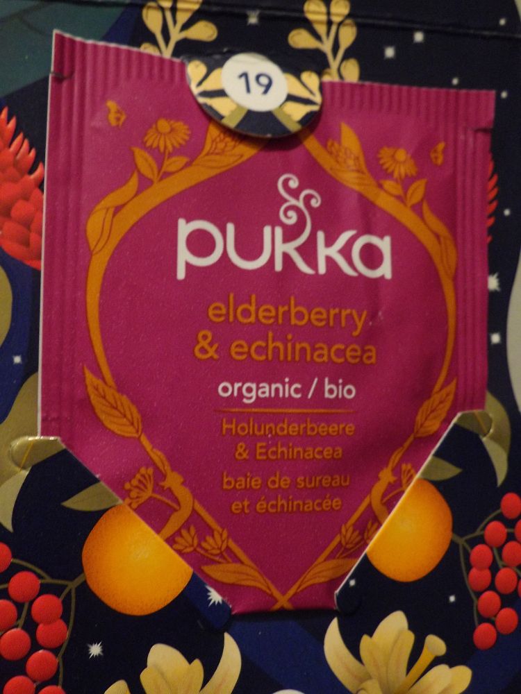 Close-up of a tea bag wrapper on a tea Advent calendar's 19th door. On a rose coloured wrapper the brand's name ('pukka') on top in white letters. Underneath the brand name the text "elderberry & echinacea" in orange letters and below that "organic/bio" in white letters. At the bottom texts "Holunderbeere & Echinacea" and "baie de sureau et échinacée" in German and French, both in orange letters. All of that surrounded by orange decoration.