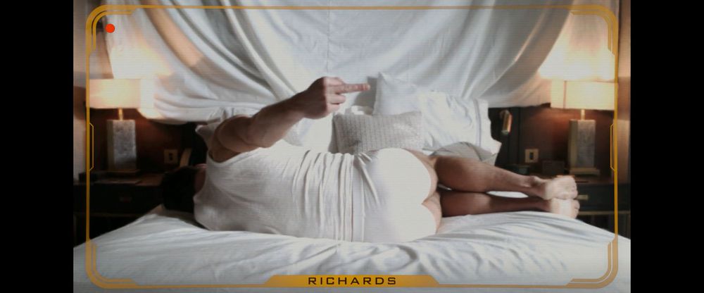 Frame from the dystopian action thriller 'The Running Man' (2025). Image through a video recording device. Yellow quadrangle with rounded corners circling the screen. Red dot on the top left corner and the name "Richards" at the bottom of the quadrangle. A man (Glen Powell as Ben Richards) wearing a white wifebeater and underpants lying sideways on his left side on a hotel room bed with his back to the camera and is giving the middle finger to the camera with his right hand. There's a white sheet covering the wall in the background.