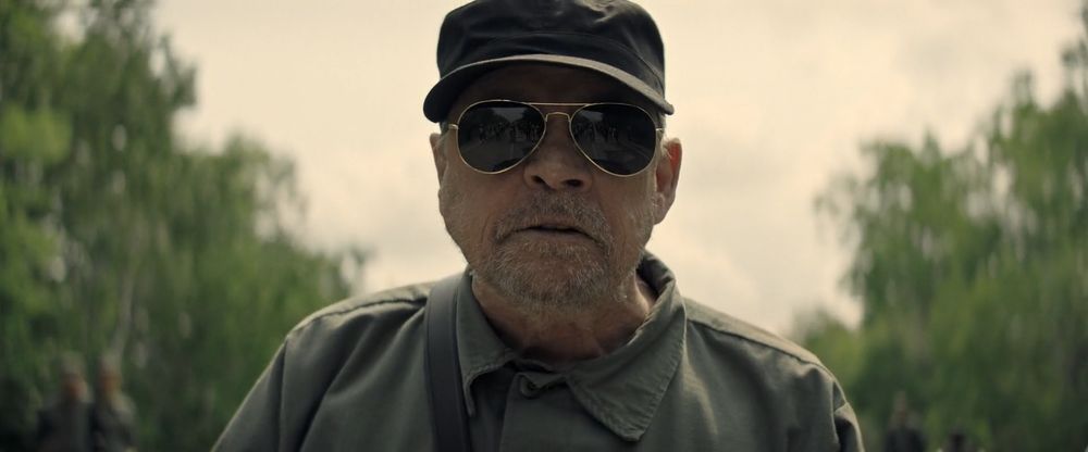 Frame from the dystopian thriller 'The Long Walk' (2025). Close-up of a older soldier (Mark Hamill as The Major) with a grey stubble wearing a green combat uniform, a green cap and sunglasses (group of young men standing on the road reflected on the lenses).