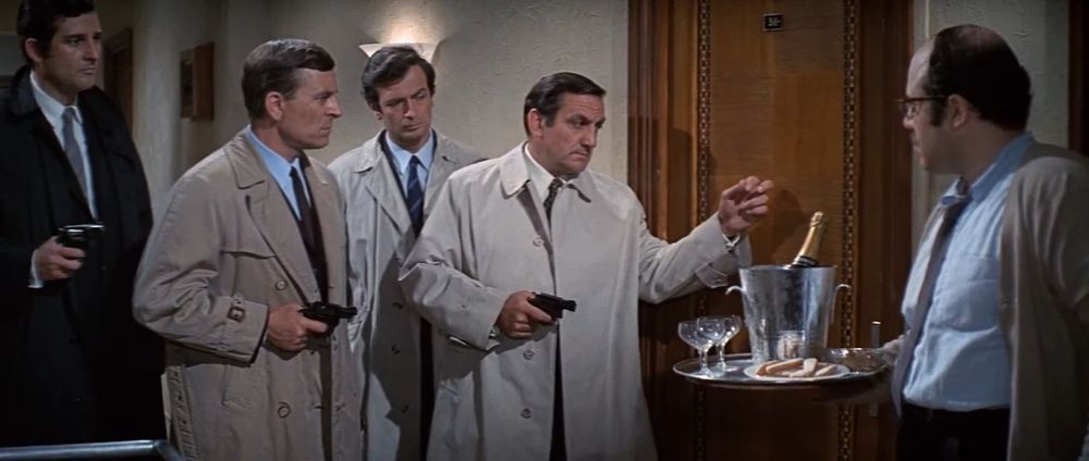 Frame from the French crime film 'Le clan des Siciliens'/'The Sicilian Clan' (1969). Four police detective wearing trenchcoats and suits (three of them holding revolvers in their right hands) standing outside a hotel room door. One of them (Lino Ventura as Chief Inspector Le Goff) is about to knock on the door. The hotel receptionist carrying a tray with champagne classes and a bucket with a champagne bottle on the right edge of the frame.