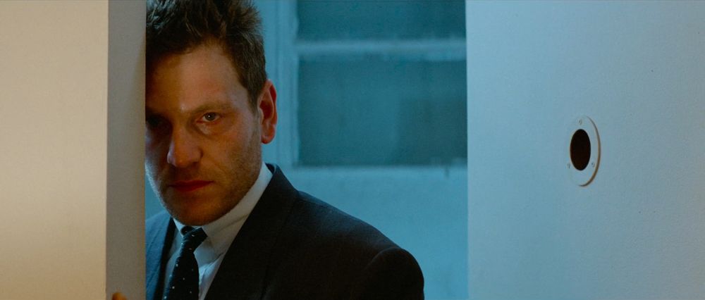 Frame from the French action thriller 'Nikita'/'La Femme Nikita' (1990). Close-up of a brown haired man (Tchéky Karyo as Bob) with stubbled beard wearing a black suit and a tie and a white shirt standing at the doorway of a room.