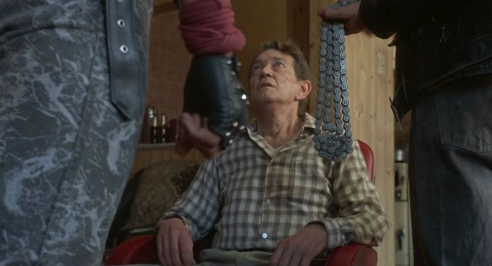 Frame from the vigilante action thriller 'Death Wish 3' (1985). A brown haired older man (Francis Drake as Charley) wearing a plaid shirt sitting in a red arm chair. Standing in front of him on both sides of the chair are two gang members (only the sides of their thighs visible on the foreground). The gang member on the right is holding a motorcycle chain in his left hand.