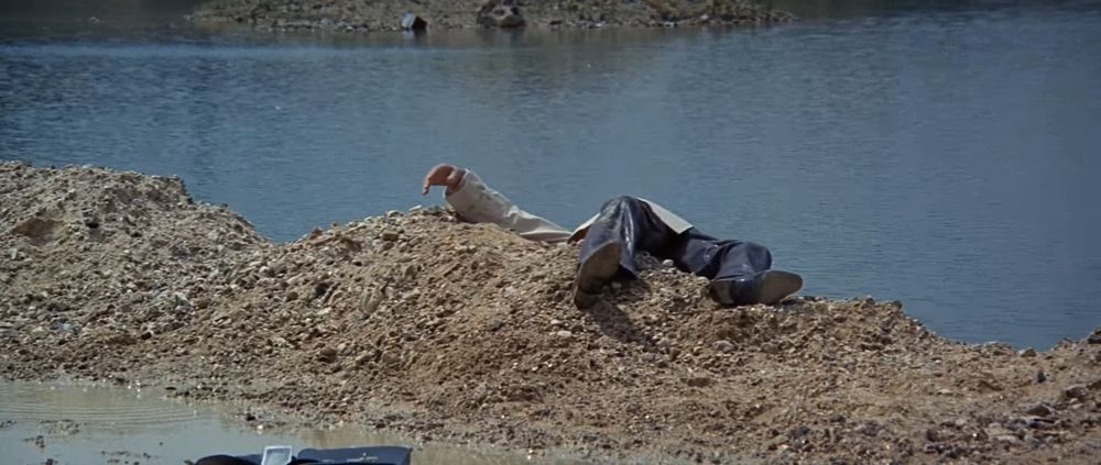 Frame from the French crime film 'Le clan des Siciliens'/'The Sicilian Clan' (1969). A dead body wearing a trench coat and dark trousers lying on a pile of a gravel in a sand quarry. Only the body's legs and right arm visible. A pool of water on the background. A black briefcase lying on the ground in the foreground.