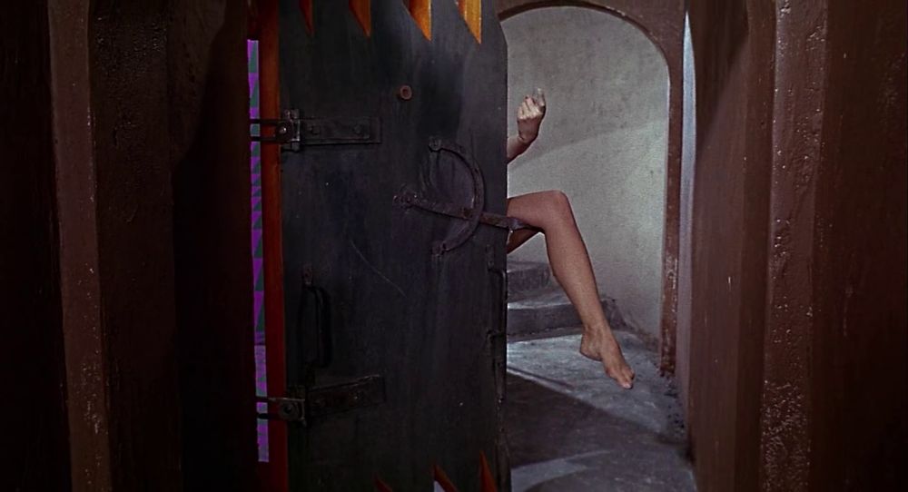 Frame from the spy comedy 'Modesty Blaise' (1966). A leg and a hand of a woman appearing from behind a partially opened steel door. The leg's resting on the latch of the door. The forefinger is making a come hither motion.
