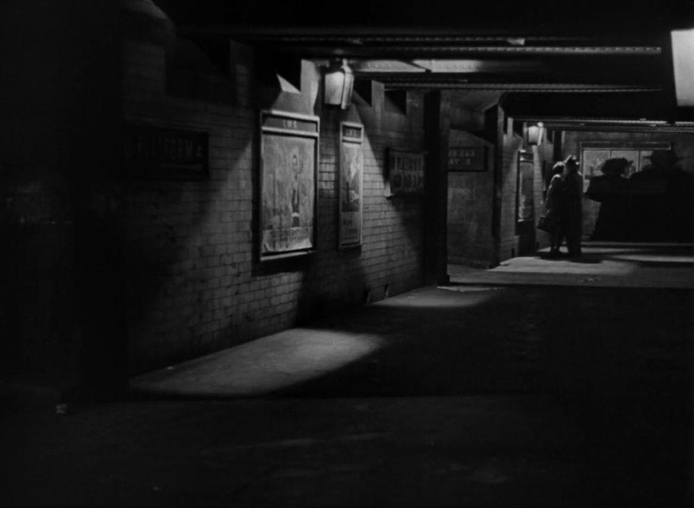 Black and white frame from the British romantic tragedy 'Brief Encounter' (1945). View of a railway station's darkly lit underpass. A woman and a man are embracing each other at the far end of the underpass. The shadow of an approaching couple visible on the wall at the end of the underpass.