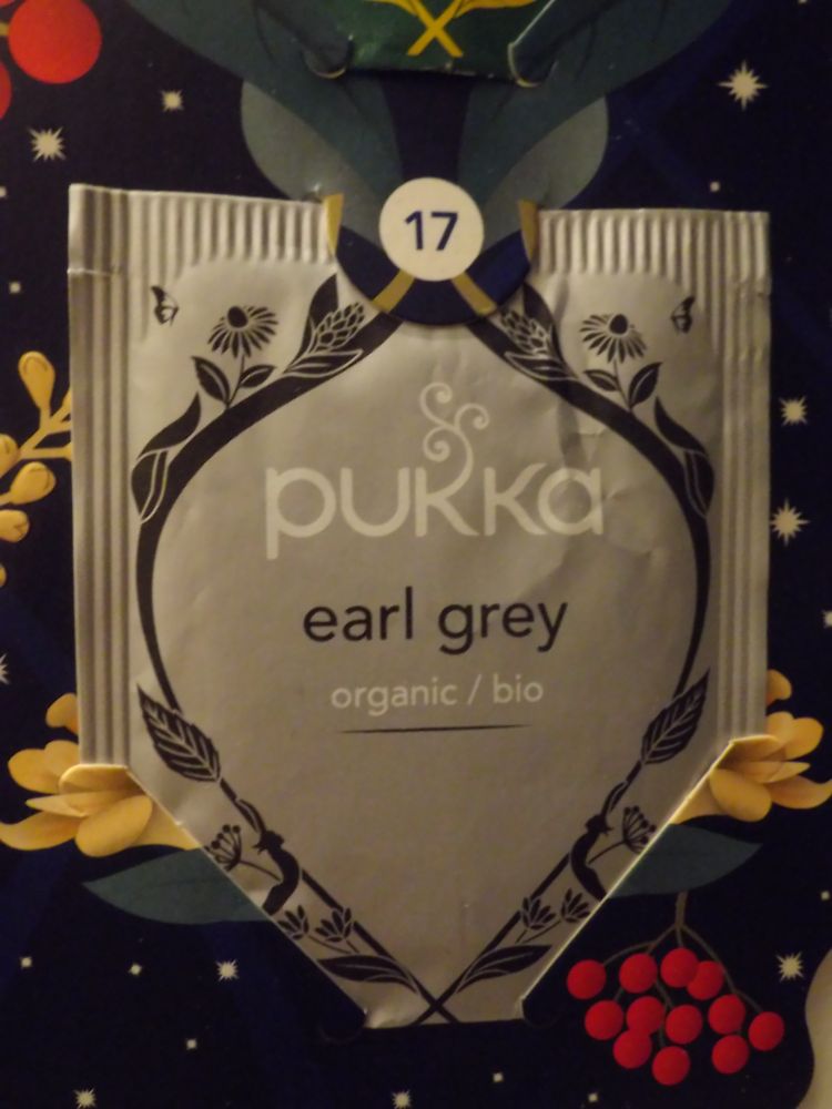 Close-up of a tea bag wrapper on a tea Advent calendar's 17th door. On a grey/silvery wrapper the brand's name ('pukka') on top in white letters. Underneath the brand name the text "earl grey" in black and below that "organic/bio" in white. All of that surrounded by black decoration.