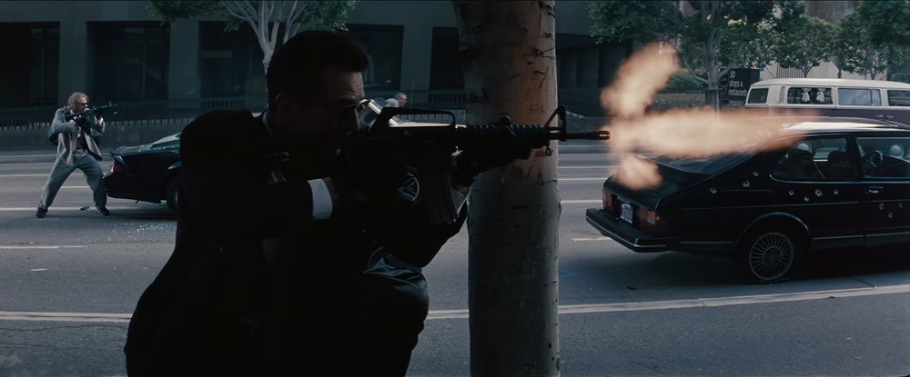 Frame from the crime film 'Heat' (1995). An armed robber (Robert De Niro as Neil McCauley) wearing a dark suit and sunglasses standing on the sidewalk next to a tree and firing a Colt Model 733 assault rifle to the right. In the background on the left another armed robber (Val Kilmer as Chris Shiherlis) in a grey suit pointing also a Colt Model 733 assault rifle to the right. Third robber (Tom Sizemore as Michael Cheritto) in the background in the middle of the frame. A black Saab with bullet holes on the street on the right with a windowed van visible behind the Saab.