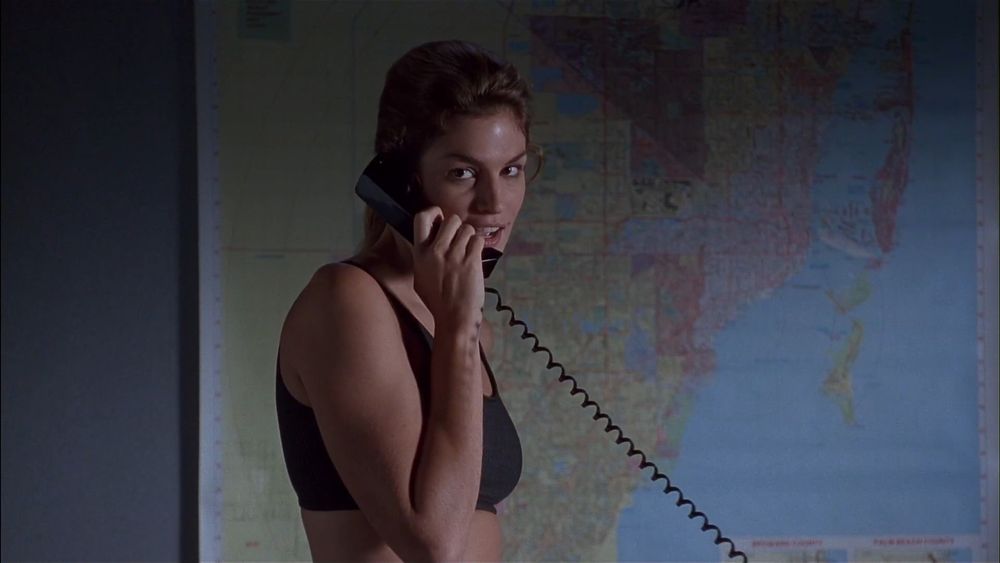 Frame from the action thriller 'Fair Game' (1995). Brown haired woman (Cindy Crawford as Kate McQuean) wearing a black sports bra standing in an office in front of a map of the Miami area talking to a telephone, she's holding the receiver to her right ear.