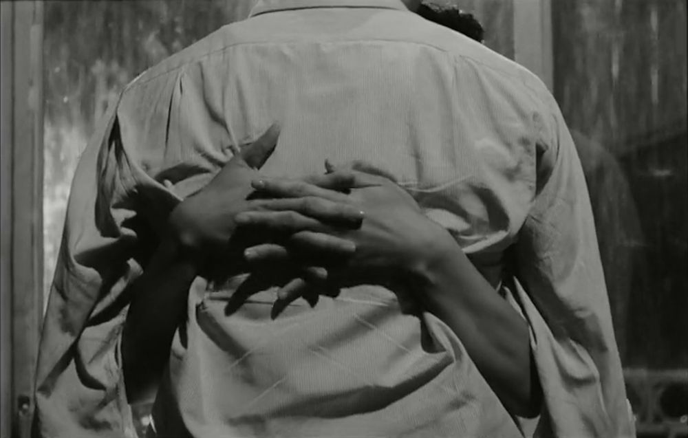 Black and white frame from the South Korean psychological thriller '하녀'(Hanyeo)/'The Housemaid' (1960). Close-up of a man's back with the arms of a woman hugging him appearing with interlocked fingers behind his back. They're standing in front of French doors at night and there's raining outside.