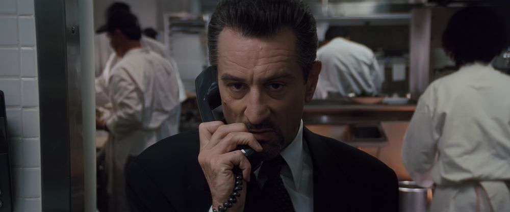Frame from the crime film 'Heat' (1995). Close-up of a dark haired goateed man (Robert De Niro as Neil McCauley) wearing a dark suit and a tie standing facing the camera in a restaurant kitchen and holding a telephone receiver in his right hand.