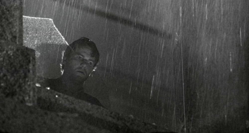Black and white frame from the neo-noir thriller 'D.O.A.' (1988). Close-up of a man (Dennis Quaid as Prof. Dexter Cornell) standing at a broken window looking down on the ground. It's raining outside.