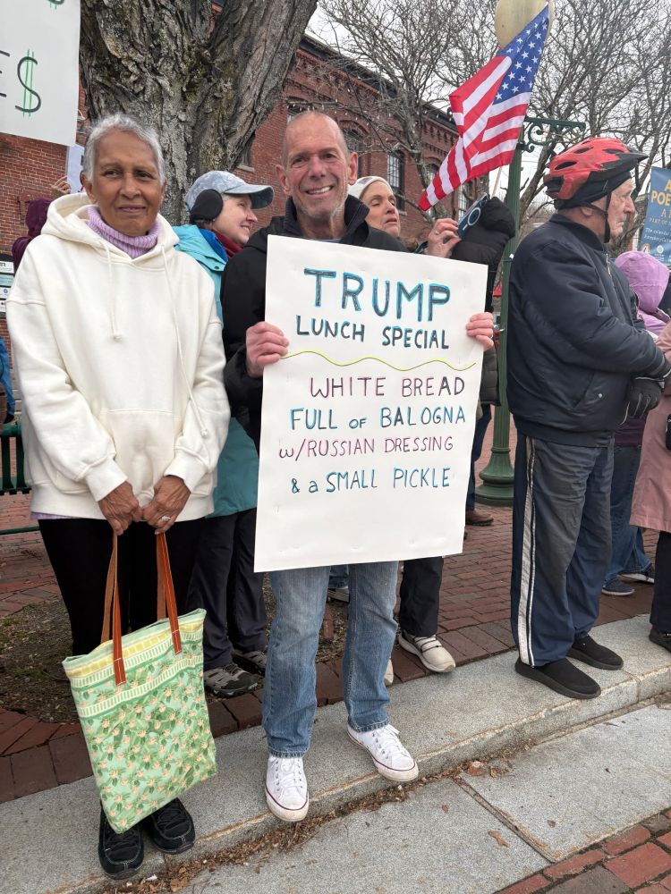 Woman and man in crowd of protestors on side of street with American flag in background. Man holds sign that reads: Trump Lunch Special. White Bread, Full of Balogna, w/ Russian Dressing & a Small Pickle.
