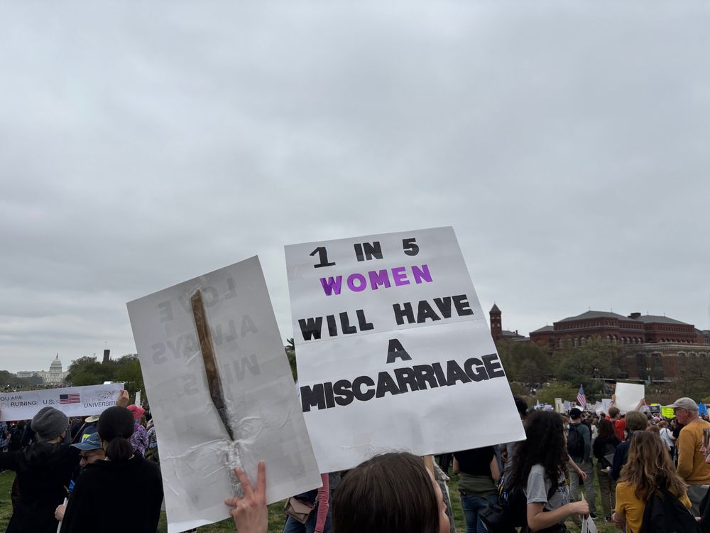 A crowd holding signs. Sign in forefront reads: "1 in 5 women will have a miscarriage" Sign is with with black lettering, and "Women" is in purple lettering"