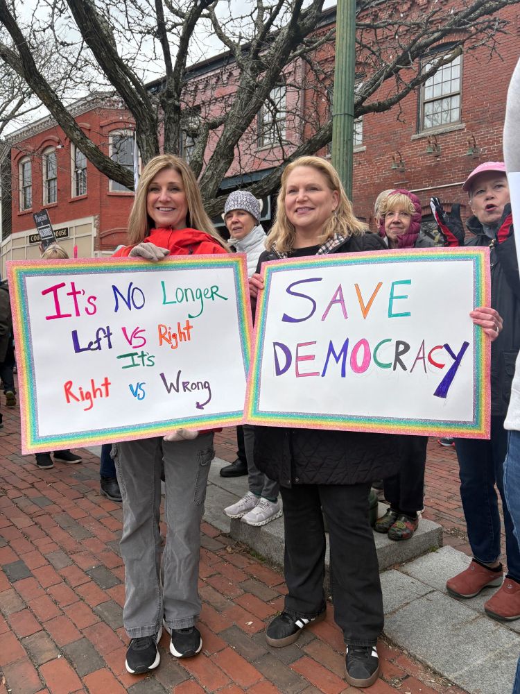 Two white women smile and pose with protest signs. Sign 1: It's no longer Left vs Right, It's Right vs Wrong. Sign 2: Save Democracy.