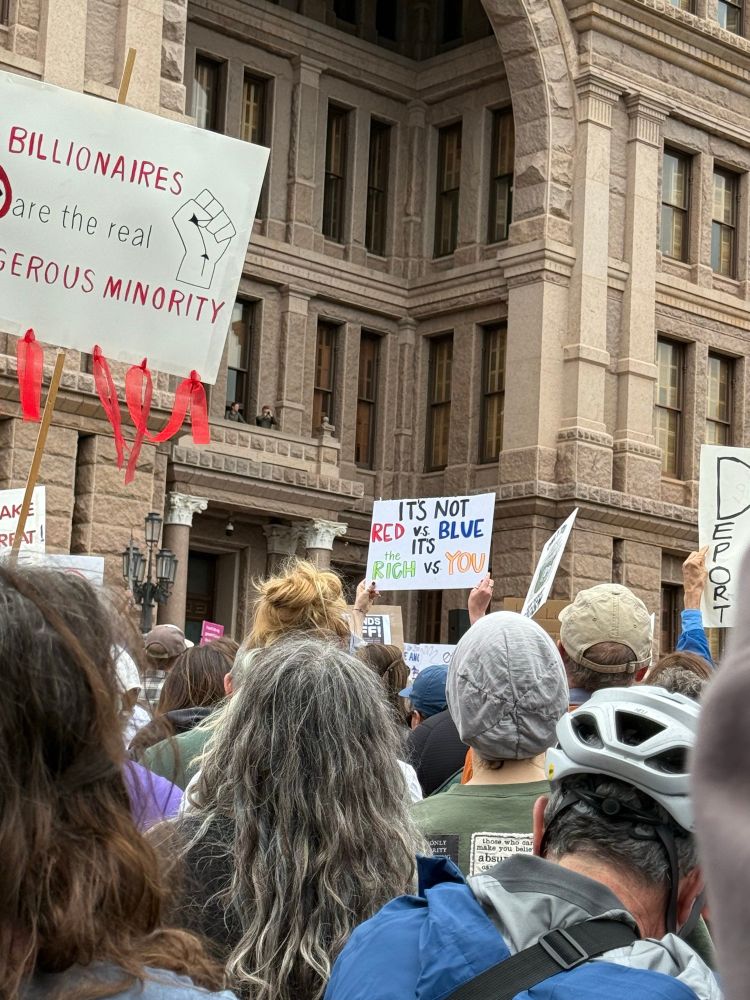 Backs of protestors heads, holding signs and facing a tan building. Sign 1: "Billionaires are the real dangerous minority" (with illustration of clenched fist). Sign 2: "It's not RED vs BLUE. It's the RICH vs YOU".