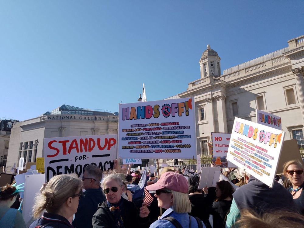 Large crowd in front of The National Gallery in London against a blue sky. Protest signs read: "Stand up for Democracy" and "Hands Off! Immigrants, Courts, Immigrants, Personal Data, Public Lands, Veteran Services, Our Wallets, Our Books, Fair Elections, Protections, Clean Air, Clean Energy, Schools, Libraries, Free Speech, LGBTQ+ Rights, Medicaid, Medicare, Social Security, Our Jobs, Cancer Research, Nato, Consumer".