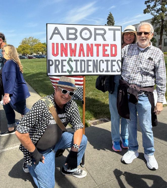 Two men and one women (all with gray hair) pose and smile next to protest sign reading "Abort Unwanted Presidencies". The S in Presidencies is a dollar sign.