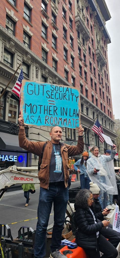 Man holding sign that reads "Gut Social Security = Mother in Law as a Roommate". Man is under a tall brick building, and wearing a brown jacket over a tee shirt with Statue of Liberty on it. Other protestors stand in background.