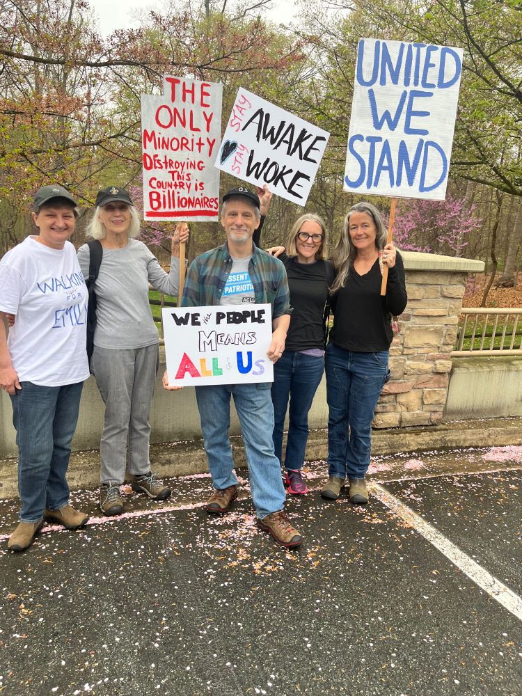 Five people posing with protest signs in front of trees and brick fence, some with arms around each other. Sign 1: "The only minority destroying this country is billionaires". Sign 2: "We the people means all of us". Sign 3: "Stay awake, Stay woke". Sign 4: "United we stand". People are standing in a parking lot with cherry blossom petals under their feet.