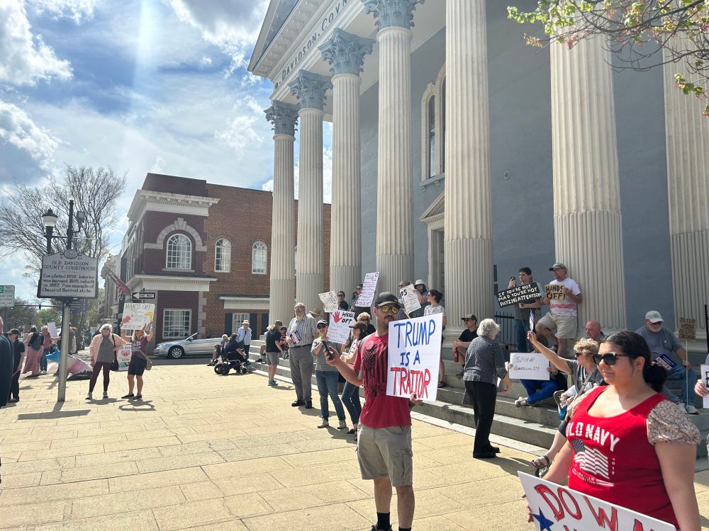 A small crowd holding protest signs in front of the columns of Old Davidson County Courthouse. Man and woman in forefront wear red shirts with American flags, and a "Trump is a Traitor" sign. Other signs in background read: Sign 1: "If you're not mad, you're not paying attention". Sign 2: "Hands off". Sign 3: "Save America". Sign 4: "So much wrong, so little cardboard". 