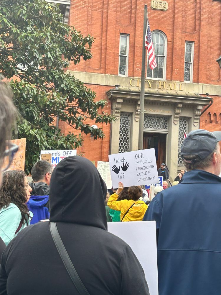 View of crowd from their backs in front of a red brick City Hall, with an American flag in front. Visible sign reads: "Hands off our schools, healthcare, bodies, democracy".