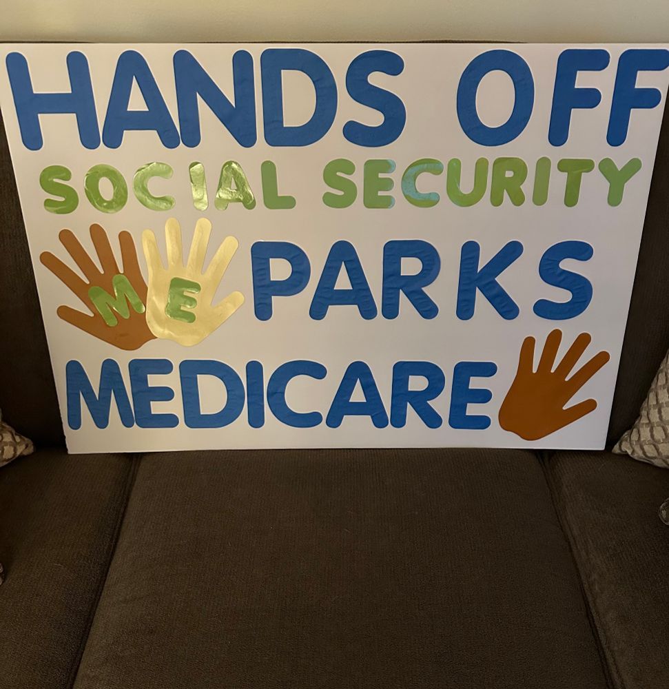 Close up protest sign on a brown couch reads "Hands off Social Security, Parks, Medicare". Brown and gold handprints decorate the sign.