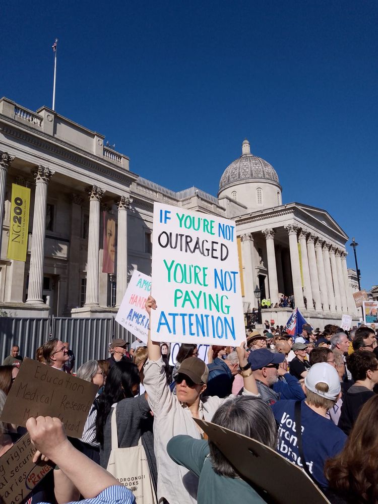Large crown in front of The National Gallery in London. Woman in forefront holds white protest sign up high that reads "If you're not outraged, You're not paying attention".