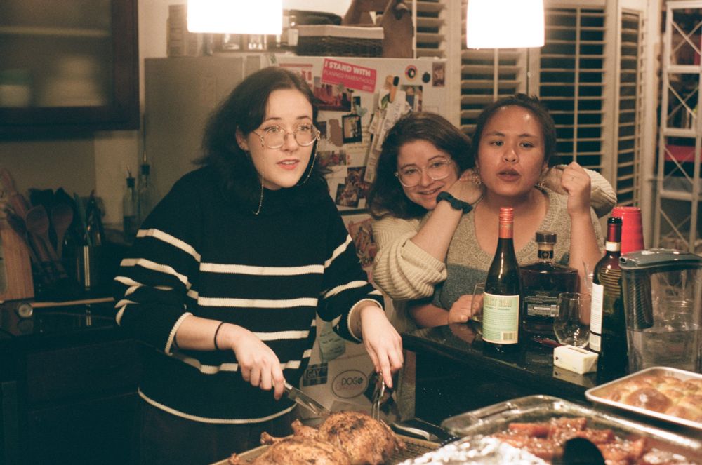 candid of three people in a kitchen; vee is carving roast chicken and talking to someone across the counter, and jess and i are standing by chatting
