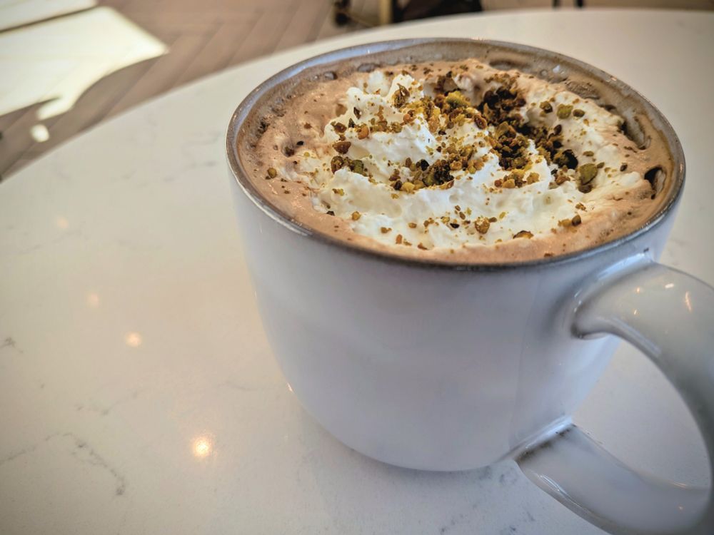 A closeup of a specialty latte in a white mug with whipped cream on top and sprinkled with pistachio pieces. 