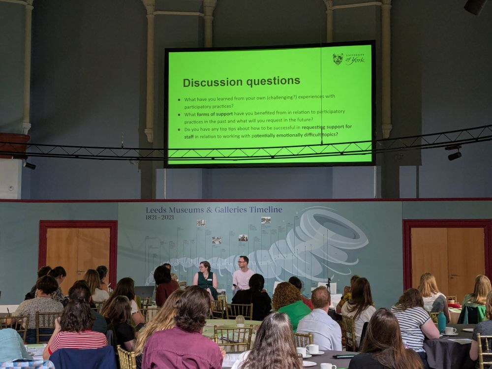 Photo shows a room of people looking at a screen. The screen is green with three discussion questions as listed in the text above 