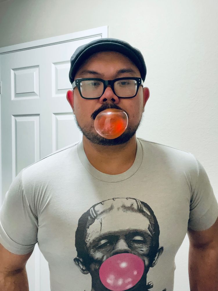 Guy wearing flat cap and glasses blowing a bubblegum bubble. He is wearing a white T-shirt with a picture of Lon Chaney’s Frankenstein’s monster blowing a bubblegum bubble.