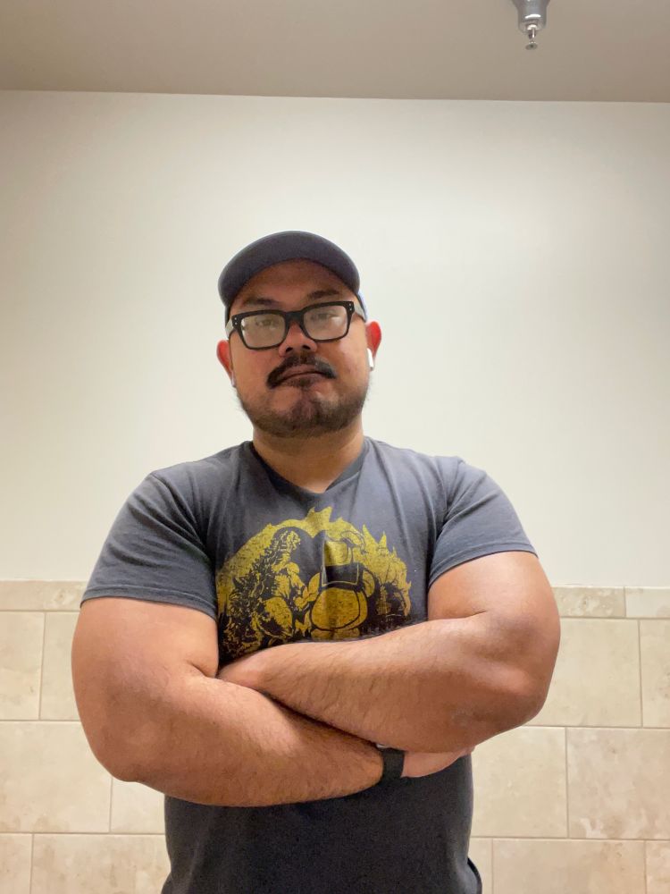 Guy in cap and black horn-rimmed glasses with arms crossed. He is wearing a black shirt with a printed image of Godzilla and the Stay Puft Marshmallow Man walking off hand in hand into the sunset.