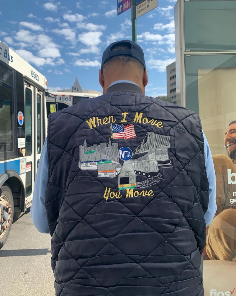 Man with his back turned to the camera wearing a navy blue quilted vest with embroidered images of trains, busses, a bridge and the city skyline. The words “when I move you move” are also embroidered in yellow