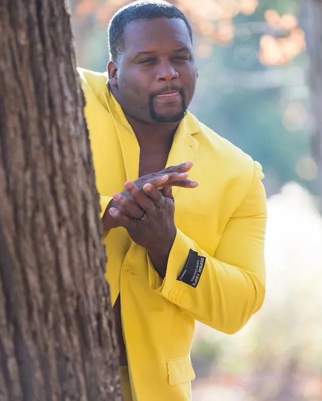 Man in yellow suit hiding behind tree, rubbing his hands together and licking his lips in anticipation