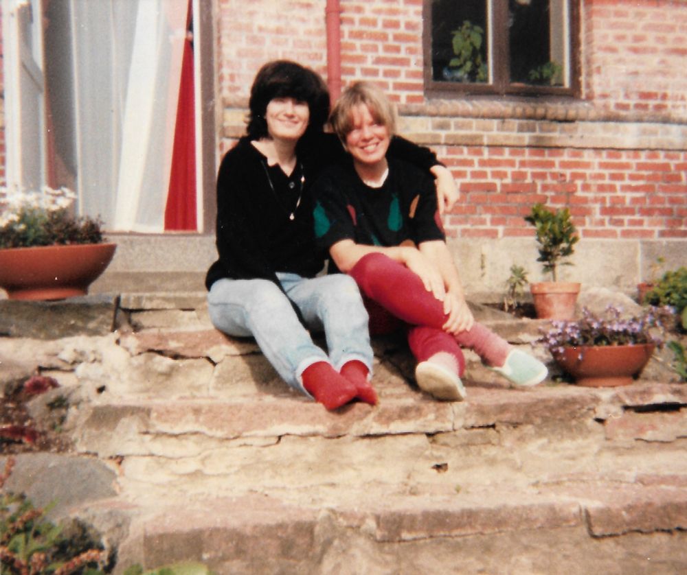 Two women sit on the steps of a red brick cottage. One woman has shoulder length dark hair. She is wearing faded blue jeans and a black sweater. She has her arm around a blonde woman who is wearing a black top with green, yellow and red print with red trousers.  There are terra cotta flower pots on a few of the steps.